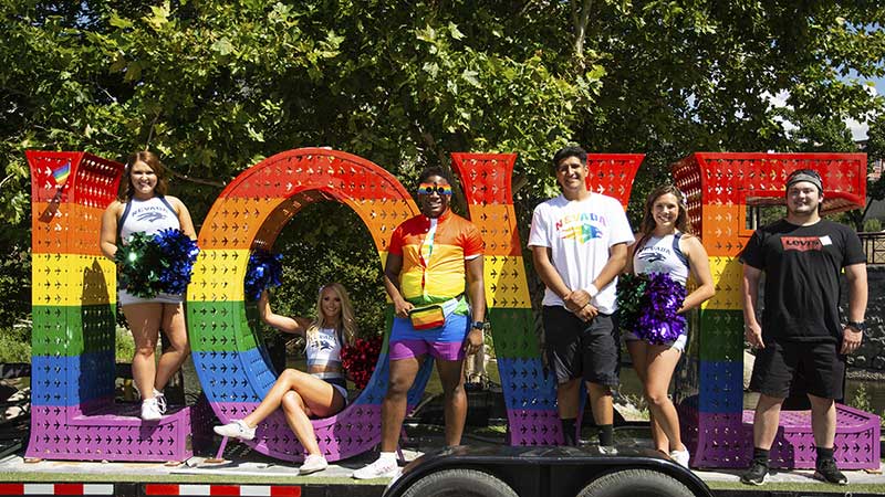A group of people, including two ϱ cheerleaders and some wearing University of ϱ, shirts, stand inside of and next to giant letters "LOVE" that are painted with the LGBTQ+ rainbow of colors, red, yellow, purple, green and indigo.