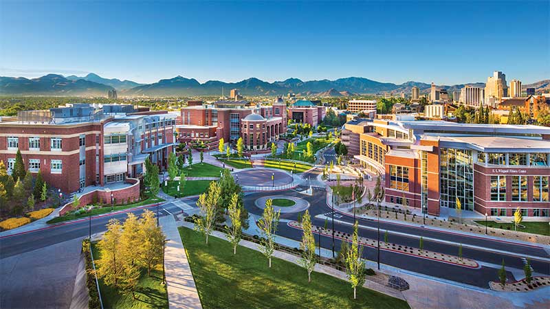 An aerial view of the University of ϱ, Reno buildings and campus looking south, showing multiple buildings, trees, and pathways. Mountains and a blue sky with clouds are visible in the distance.